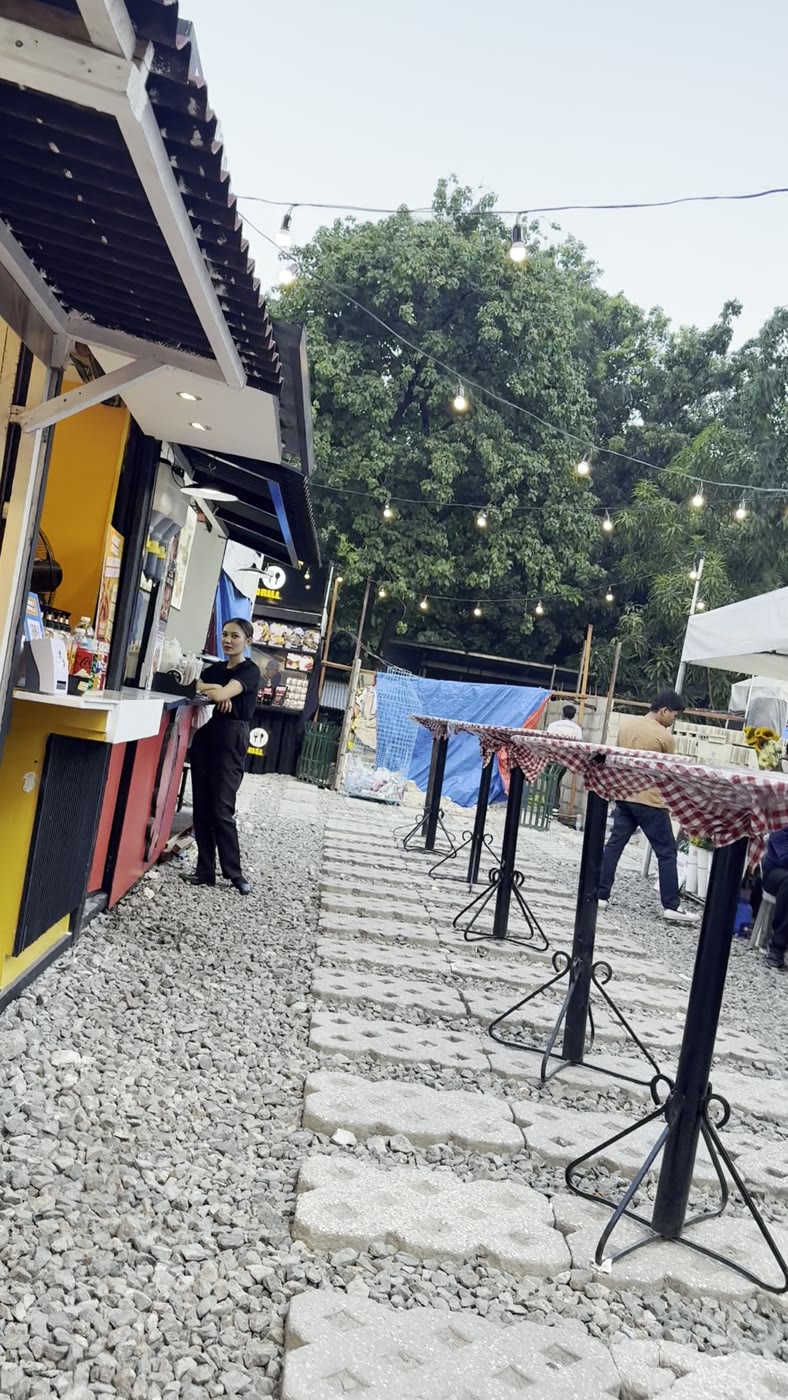 砂利とコンクリートブロックの新しい会場に並ぶ立ち飲みテーブルと電球のガーランド